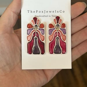 May include: A pair of colorful earrings with a unique design. The earrings feature a rectangular shape with a floral-like pattern in shades of red, pink, purple, and orange. The text "The Fox Jewels Co Handcrafted in Texas" is visible on the card.