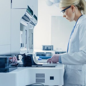 以下が含まれることがあります： 白衣を着た女性が、実験室で医療機器を操作しています。機器は白で、青いディスプレイが付いています。