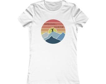 Puede incluir: Camiseta blanca con un diseño gráfico circular que representa un paisaje montañoso y una persona saltando. El diseño gráfico incluye un diseño de puesta de sol y el texto "Don't Be a Victim... Be a VICTOR!"