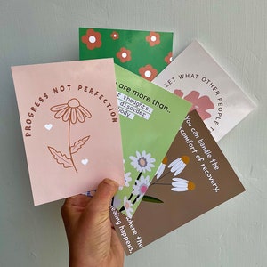 May include: Four cards with floral designs and inspirational messages. The cards are pink, green, brown, and white with brown text. The messages on the cards are "Progress not perfection", "You can handle the discomfort of recovery", "Let what other people think", and "You are more than your thoughts, disorder, body."