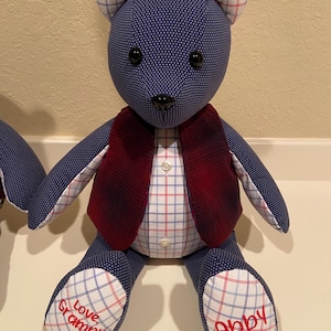 May include: A handmade teddy bear with a navy blue polka dot body, plaid ears, and a red plaid vest. The bear has black button eyes and a black nose. The bear's feet are white with a red and blue plaid pattern, with the names "Love Grampy" and "Abby" embroidered in red.