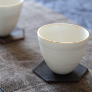 Puede incluir: Dos tazas de té de cerámica blanca con un borde ligeramente ensanchado sobre posavasos hexagonales marrón oscuro. Las tazas están sobre una superficie textil gris, con un textil azul en el fondo. Diseño minimalista.