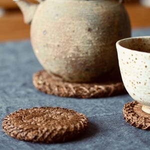 Puede incluir: Un juego de té rústico con una tetera de cerámica texturizada y una taza de té blanca moteada, sobre posavasos marrones tejidos. Los posavasos redondos tienen un borde trenzado. El juego está sobre una superficie azul.