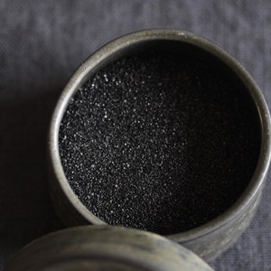 May include: A round, metal container with a lid. The container is filled with black, granular material.