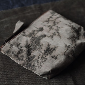 Könnte beinhalten: Eine kleine, graue Tasche mit einem schwarz-weißen Landschaftsdesign. Die Tasche hat eine rechteckige Form mit einer kleinen Schlaufe an der Seite. Der Hintergrund ist ein dunkelgrauer Stoff.