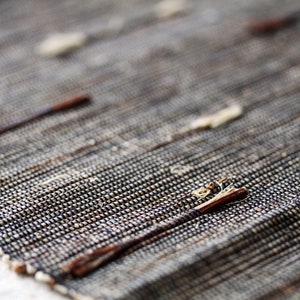 以下が含まれることがあります： 織り込まれた質感のある表面を持つ敷物のクローズアップ。敷物は天然繊維で作られており、微妙な茶色と黒の色合いがあります。