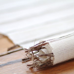 May include: A white linen table runner with a subtle striped pattern is rolled up and tied with a brown twig. The runner is lying on a wooden surface.