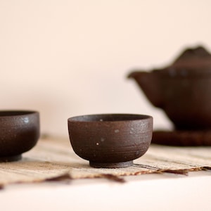 May include: Two small brown ceramic teacups and a larger brown ceramic teapot on a woven mat. The cups and teapot have a rough, textured surface.