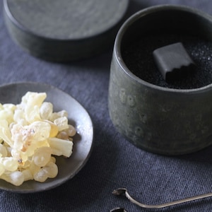 Puede incluir: Arreglo de elementos para quemar incienso. Incluye un plato pequeño con trozos de resina amarillo pálido, una taza de cerámica gris oscuro con una sustancia negra y un bloque gris oscuro, un plato pequeño y pinzas plateadas.