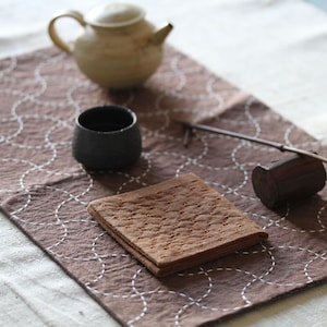 Könnte beinhalten: Ein braunes Stofftischset mit einem weißen gestickten Muster aus Kreisen. Eine kleine braune Holzkiste, eine Keramik-Teetasse und eine Keramik-Teekanne befinden sich auf dem Tischset.
