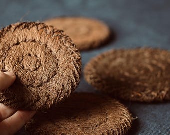 May include: Close-up of several round, brown coasters made from woven natural fibres. One coaster is being held by a hand. The coasters have a spiral design in the centre and are placed on a dark blue surface.