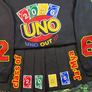 May include: Black sweatshirt and pleated skirt set featuring a colorful UNO card game design. The sweatshirt displays the word "UNO" and the numbers "2026". The skirt has the text "class of" and "Ja'Mya" with UNO card graphics.