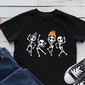May include: Black t-shirt with a graphic of four skeletons dancing. The skeletons are wearing hats and holding pumpkins. The shirt is on a white wooden surface with a pair of blue jeans and white sneakers.