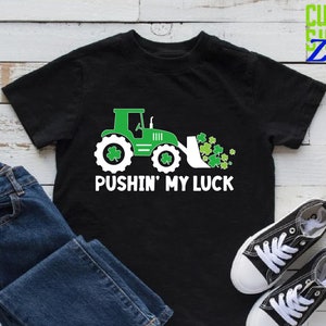 May include: A black t-shirt with a green tractor pushing a pile of shamrocks. The text "Pushin' My Luck" is printed in white below the tractor.