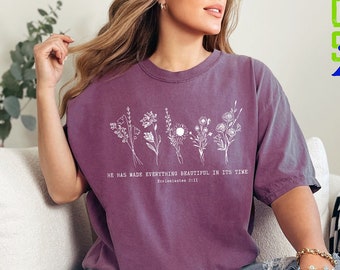 Puede incluir: Una camiseta morada con un diseño floral blanco y el texto "Él ha hecho todo hermoso en su tiempo" con la referencia bíblica "Eclesiastés 3:11".