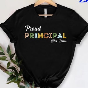 May include: A black short-sleeved t-shirt featuring the white text Proud and the word PRINCIPAL in multi-colored block letters, followed by the white text Mrs Davis. The shirt is displayed on a wooden hanger with a green leafy branch underneath.