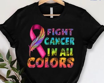 Puede incluir: Camiseta negra con una cinta rosa, una pluma arcoíris y el texto "Fight Cancer in All Colors".