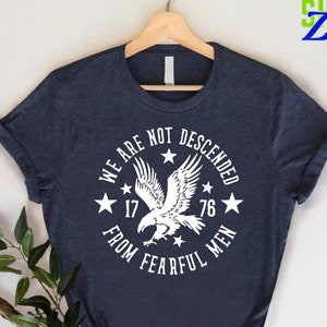 May include: A navy blue t-shirt with a white graphic of an eagle and stars. The text on the shirt reads "We are not descended from fearful men 17 76".