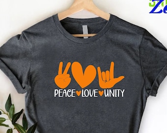 Pode incluir: Uma t-shirt cinza-carvão escuro com um gráfico laranja de um sinal de paz, um coração e um gesto de mão de eu te amo. Abaixo do gráfico, o texto branco diz PEACE LOVE UNITY, separado por pequenos corações laranja num cabide.