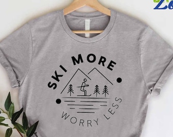 以下が含まれることがあります： グレーのTシャツに、黒いグラフィックデザインが施されています。「Ski More Worry Less」の文字と、山の中でスキーをしている人のイラストが描かれています。