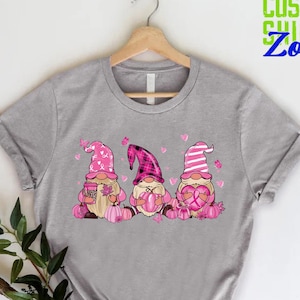 May include: A gray t-shirt featuring three gnomes with pink hats and pumpkins. The gnomes are holding a coffee cup, a pumpkin, and a heart. The text "CUSTOM SHIRTS Zone" is in green and blue.