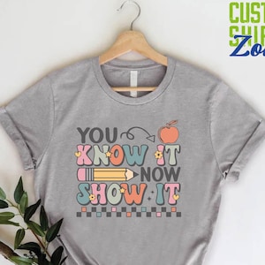 May include: A gray t-shirt with the text "YOU KNOW IT NOW SHOW IT" in colorful, retro-style lettering. The text is accompanied by a pencil, an apple, and a checkered pattern.