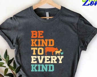 May include: A dark grey t-shirt with the text "Be Kind To Every Kind" in a retro color scheme. The text is stacked vertically with a cow, goat, and chicken graphic between the words "To" and "Every".