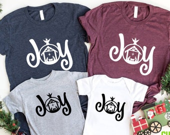 May include: A set of four Christmas shirts featuring the word "Joy" with a manger scene inside a circle. The shirts are in various colors, including navy blue, maroon, gray, and white.
