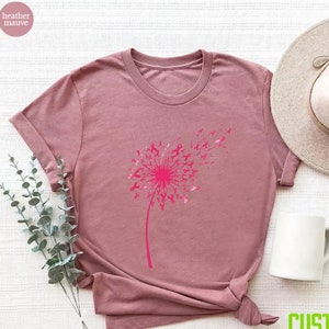 Op de afbeelding: Een heide-paarse t-shirt met een roze paardenbloemontwerp. De paardenbloem is gemaakt van roze linten, ter symbolisering van borstkankerbewustzijn.