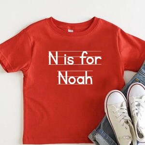 May include: Red short-sleeved t-shirt with white text that says "N is for Noah" on a white lined background.
