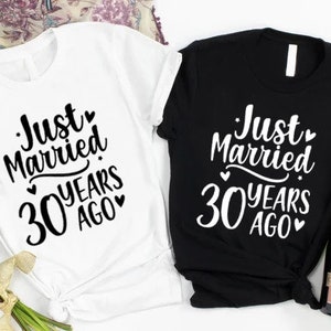 May include: Two t-shirts, one white and one black, with the text 'Just Married 30 Years Ago' printed on them. The shirts are laid out on a white surface with a bouquet of pink roses and a bottle of champagne.