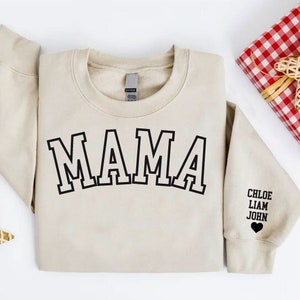Puede incluir: Una sudadera beige con la palabra "MAMA" impresa en negro en la parte delantera. La sudadera tiene un coraz&oacute;n negro con los nombres "CHLOE, LIAM, JOHN" impresos en la manga.