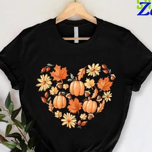 May include: Black t-shirt with a heart-shaped design made of fall leaves, pumpkins, acorns, and flowers. The design is in shades of orange, yellow, and brown.