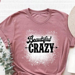Puede incluir: Una camiseta de color malva con un dise&ntilde;o de salpicaduras blancas y el texto "Beautiful Crazy" en negro.