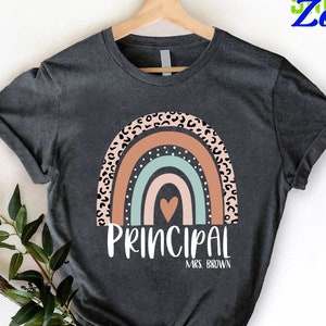 May include: A dark gray short-sleeve t-shirt featuring a graphic of a rainbow with a leopard print outer arch, a solid peach arch, and a light blue inner arch with white dots. A small heart is centered, with the text PRINCIPAL MRS. BROWN below.