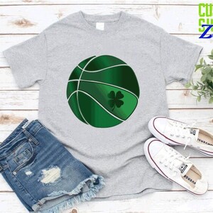 Puede incluir: Una camiseta gris con una pelota de baloncesto verde con un dise&ntilde;o de tr&eacute;bol de cuatro hojas. La camiseta est&aacute; sobre una superficie de madera blanca con una planta verde y zapatillas blancas.