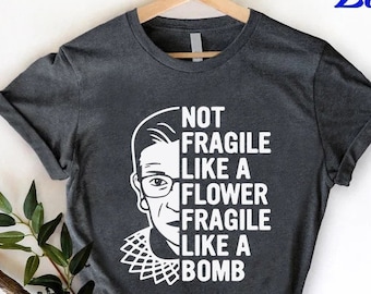 Może przedstawiać: Szary t-shirt z białym obrazkiem Ruth Bader Ginsburg i tekstem "Not Fragile Like A Flower Fragile Like A Bomb".