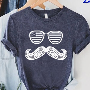 May include: Navy blue t-shirt featuring a white graphic of sunglasses with an American flag design and a large mustache. The text "CUSTOM SHIRTS Zone" is in the upper right corner. The shirt is hanging on a wooden hanger.