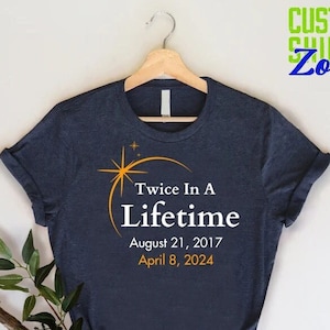 May include: A navy blue t-shirt with white text that reads "Twice In A Lifetime" with the dates "August 21, 2017" and "April 8, 2024" underneath. The shirt is hanging on a wooden hanger.