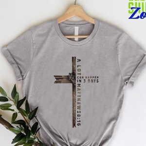May include: A gray t-shirt with a distressed wood cross design. The cross has the text "A LOT CAN HAPPEN IN 3 DAYS MATTHEW 28:16" printed on it.