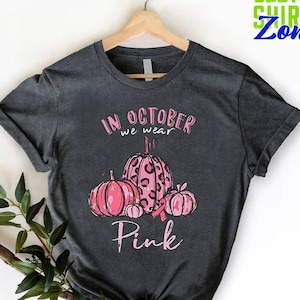 May include: A dark gray t-shirt with a pink and black pumpkin design. The text on the shirt reads "In October we wear Pink".