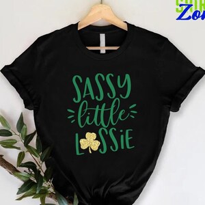 K&ouml;nnte beinhalten: Schwarzes T-Shirt mit gr&uuml;nem Text, der "Sassy little Lassie" lautet, mit einem goldenen Glitzer-Kleeblatt.