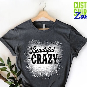 Puede incluir: Una camiseta gris oscuro con un dise&ntilde;o de pintura en aerosol blanca. El dise&ntilde;o presenta el texto "Beautiful CRAZY" en negro.
