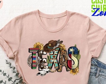 May include: A pink t-shirt with a graphic design of the word "TEXAS" in colorful letters. The letters are decorated with various western-themed elements, including a cowboy hat, sunflowers, a horseshoe, and a cactus.