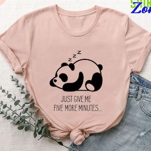 May include: A heather peach-colored t-shirt featuring a sleeping panda bear graphic in black and white. The text "JUST GIVE ME FIVE MORE MINUTES..." is printed below the panda. The shirt has a crew neck and short sleeves.