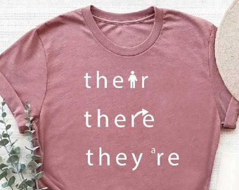 May include: A heather mauve t-shirt with the words "their", "there", and "they're" printed on it. The word "there" has an arrow pointing to the right.