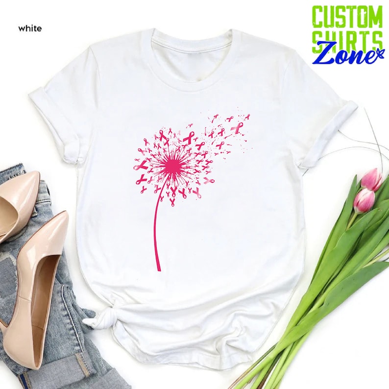 Op de afbeelding: Wit T-shirt met een roze paardenbloemontwerp gemaakt van roze linten. De paardenbloem waait in de wind.