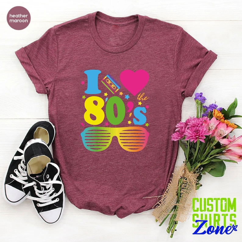 May include: Heather maroon t-shirt with a graphic design that says "I love the 80's" with a cassette tape, heart, and sunglasses.