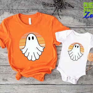 May include: Orange and white t-shirt with a ghost in a sunset design.  The design features a white ghost with a face and a sunset with orange, yellow, and brown stripes.  The t-shirt is paired with a white baby onesie with the same ghost and sunset design.
