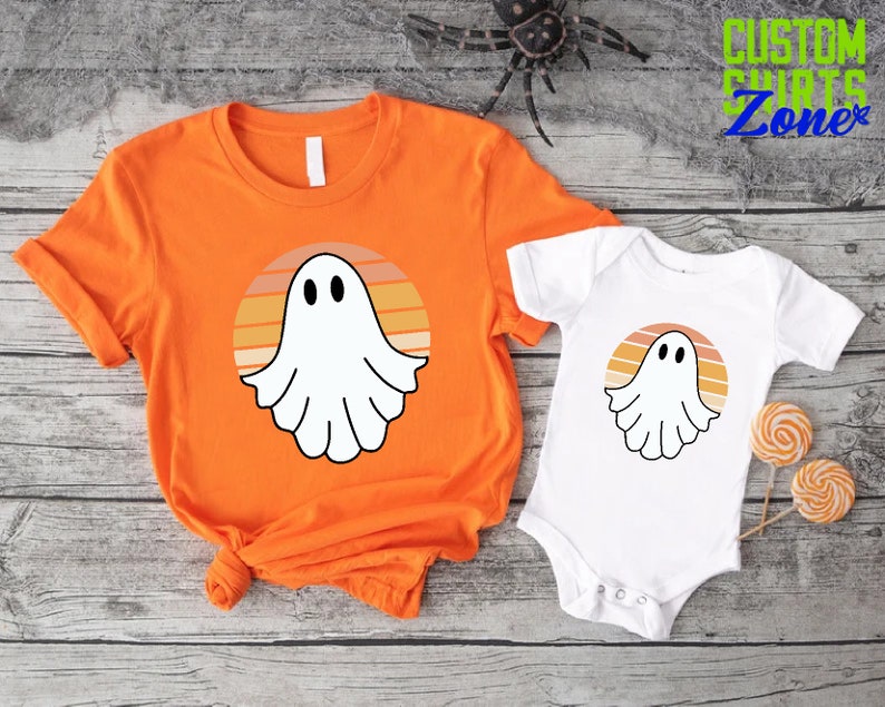 May include: Orange and white t-shirt with a ghost in a sunset design.  The design features a white ghost with a face and a sunset with orange, yellow, and brown stripes.  The t-shirt is paired with a white baby onesie with the same ghost and sunset design.